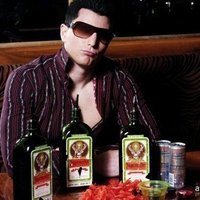Guy in sunglasses and an open shirt sitting behind bottles of Jaegermeister bottles and Redbull cans