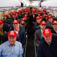 A photograph of the trump team on a plane with Ben Carson seated in the center aisle. All of the subjects are wearing keep iowa great