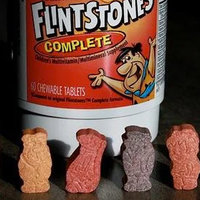 Six multi-colored vitamins shaped like characters from the Flintstones cartoon series standing in front of a bottle of Flintstones complete vitamins. 