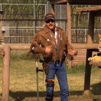 Joe Exotic in a fringe jacket leaning on a cane near a tiger reclining on a wooden platform