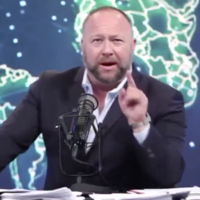Alex Jones in a blazer speaking into a mic in front of a backdrop of the world map