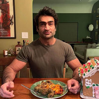 Kumail Nanjiani seated at a dinner table behind a plate of food with a gingerbread house on his left. 