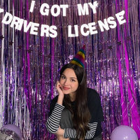 Olivia Rodrigo in a party hat in front of a glittery purple backdrop and a sign that reads "I got my drivers license"