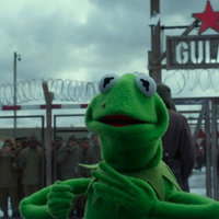 Kermit in the Gulag a screenshot from the film "muppets most wanted." In the foreground, kermit the frog, a green frog muppet, stands with his arms around his chest. In the background, there is an iron gate that reads "gulag"