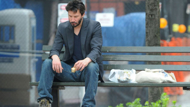 Sad Keanu Reeves sitting on a public bench and half-heartedly eating a sandwich / sad keanu meme.