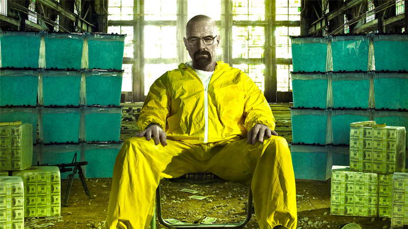 Breaking Bad | Photo of Walter White, played by Bryan Cranston, sitting among organized totes of blue meth and neatly wrapped massive cubes of cash money, as he sits tensely in a low folding chair while wearing a full body yellow hazmat suit