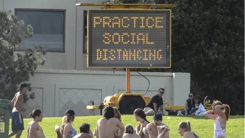 An image of people gathering in a massive group in front of a social distancing sign.