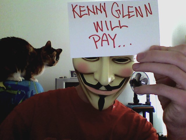 man wearing an anonymous mask and holding sign Kenny Glenn will pay and a cat in the background