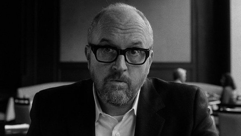 A black and white headshot of Louis CK wearing thick black framed glasses.