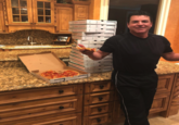 John Schnatter dressed in black standing in a kitchen in front of a tower of pizza boxes and holding a slice in one hand