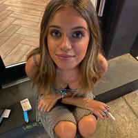 Model Brooke Flecca looking up into the camera while sitting on the curb outside of a building, smoking a c--------. Her c-------- pack lies beside her.