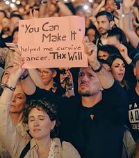 A screenshot from Will Smith's alleged AI crowd video. The audience member holds up a sign claiming Will Smith's song "You Can Make It," released in 2025, helped him survive cancer.