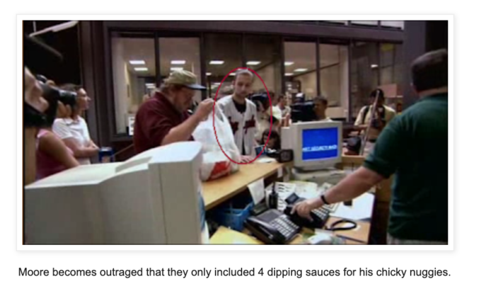 Michael Moore looking inside a bag in a busy office setting. Moore becomes outraged that they only included 4 dipping sauces for his chicky nuggies.