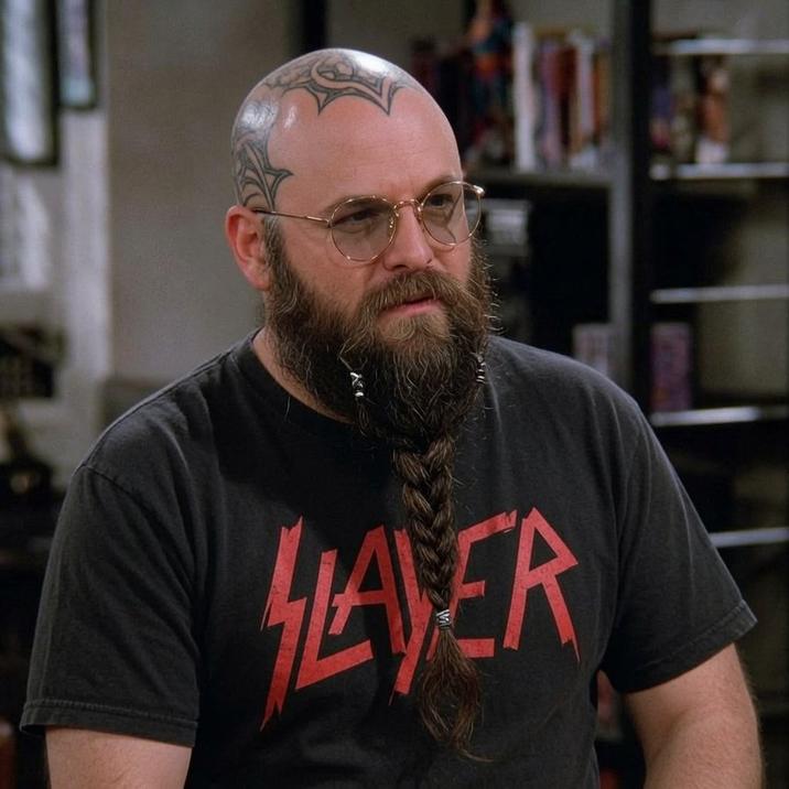 George Costanza in a Slayer shirt with a braided beard. He also has head tattoos.