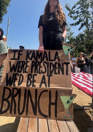 A photo of a woman holding a sign at a June 2025 "No Kings" protest, which reads, "If "Kamala":https://knowyourmeme.com/memes/people/kamala-harris were president, we'd be at brunch."