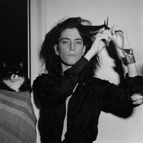 Patti Smith holding a pair of scissors to her hair, with a tuxedo cat sitting beside her, captured i...